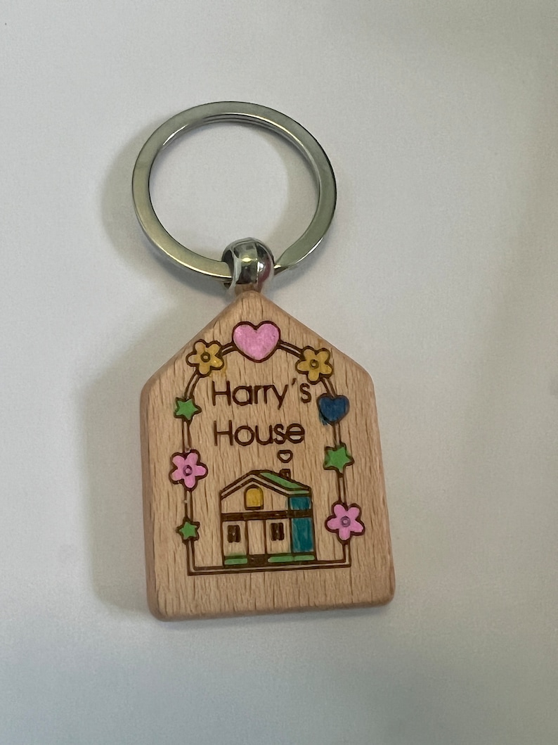 May include: Wooden house-shaped keychain with a light brown finish. The keychain features a small house with a green roof, yellow windows, and a blue door. The text "Harry's House" is engraved on the keychain, surrounded by flowers and hearts.