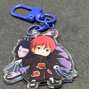May include: An acrylic keychain featuring a chibi-style character with red hair wearing a black cloak with red cloud patterns. The character is accompanied by a purple-skinned figure and holds blue strings extending from their hands to the figure.