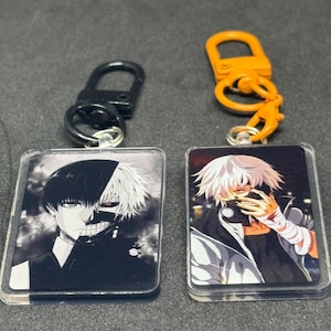 May include: Two rectangular keychains featuring anime character art. One keychain has a black and white image of a character with half of their face visible. The other keychain has a character with white hair and bandages. Each keychain has a metal clasp.