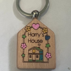 May include: Wooden keychain with a house design, a heart, and the text "Harry's House". The house is blue, green, and yellow. The keychain has a silver ring.