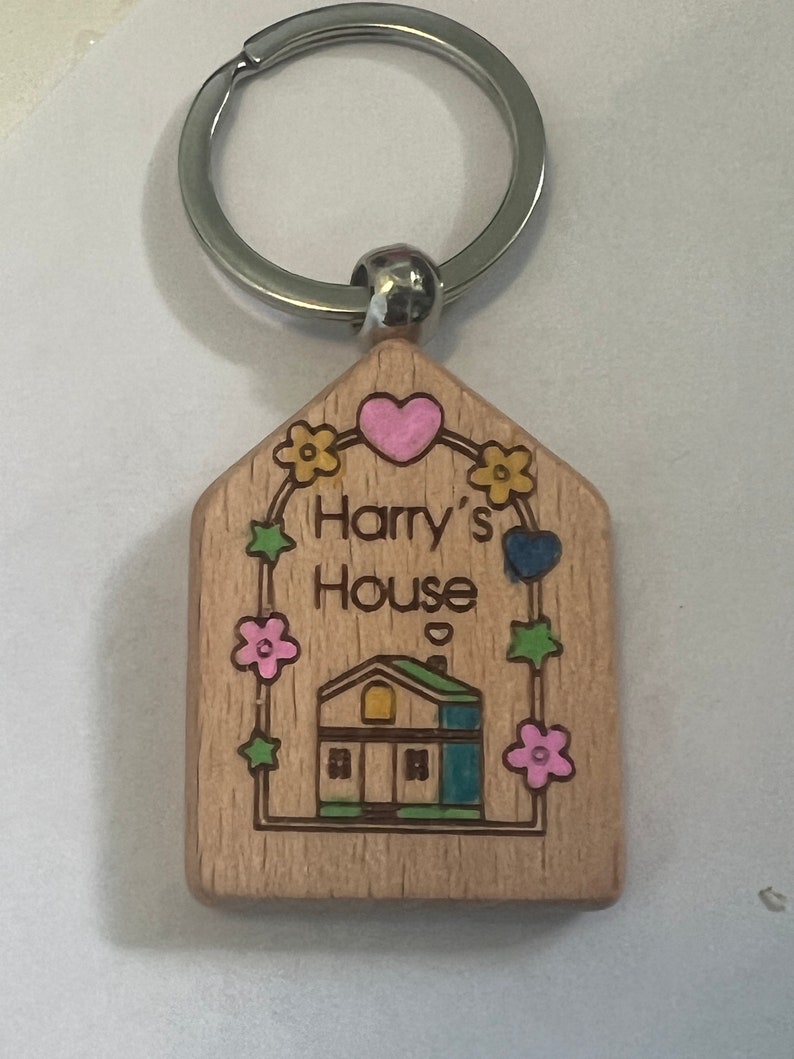 May include: Wooden keychain with a house design, a heart, and the text "Harry's House". The house is blue, green, and yellow. The keychain has a silver ring.