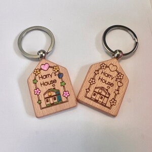 May include: Two wooden house-shaped keychains with silver key rings. Both keychains feature a small house with a green roof, a pink heart, and the text "Harry's House".