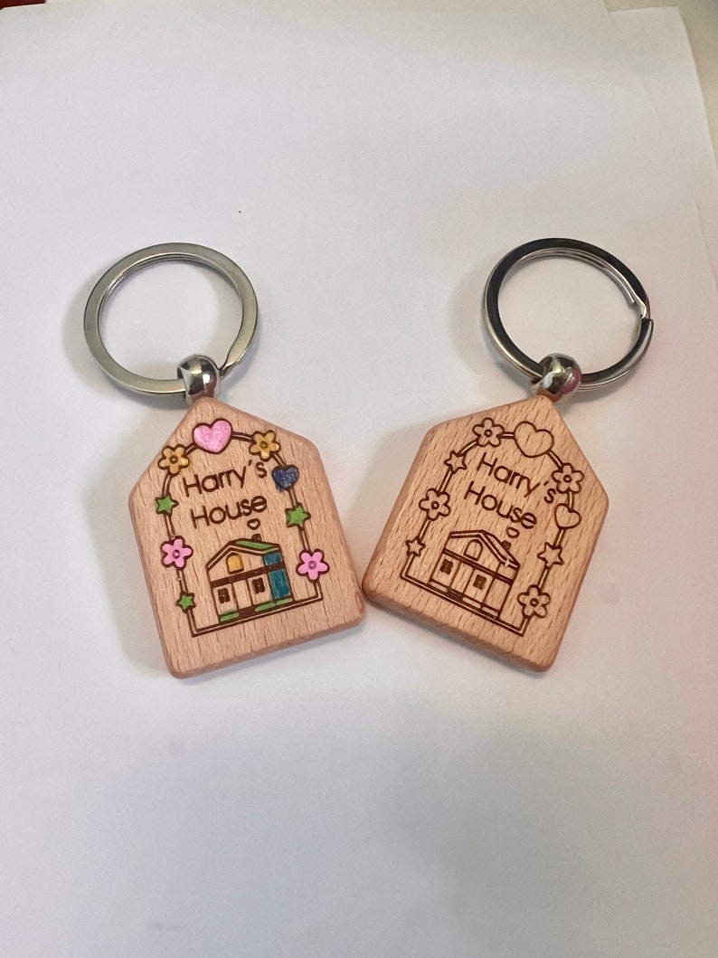 May include: Two wooden house-shaped keychains with silver key rings. Both keychains feature a small house with a green roof, a pink heart, and the text "Harry's House".