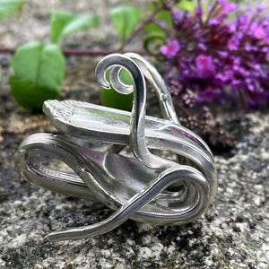 May include: A silver-colored metal ring crafted from a bent spoon, featuring a polished finish and intricate decorative handle patterns that curve into a circular band shape, showcasing the original embossed details of the silverware.