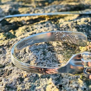 May include: A silver bracelet made from a repurposed fork, with decorative details on the handle. A spoon is visible in the background. The bracelet is resting on a textured, gray and brown surface.