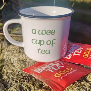 May include: A white ceramic mug with a dark rim featuring the green text "a wee cup of tea" printed on the front. Two red rectangular tea bags labeled "Barry's Tea Gold" are positioned in front of the mug on a textured stone surface.