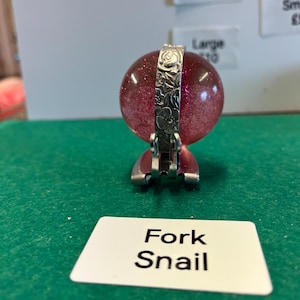 Puede incluir: Un tenedor decorativo para caracoles. El tenedor tiene una banda de metal plateado con detalles florales alrededor de un orbe rosa brillante. El tenedor está sobre una superficie verde con una etiqueta blanca que dice "Fork Snail".