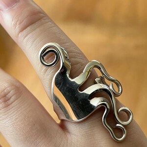 May include: A silver-toned ring crafted from a repurposed metal fork, featuring a wide, polished band that transitions into curved, spiraling tines that extend outward, creating an intricate, fluid, and sculptural design on the finger.