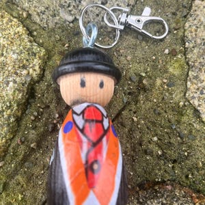 May include: A wooden figurine keychain with a black top hat, a black suit, a red tie, and a white shirt. The figurine is painted with orange, black, and blue details.
