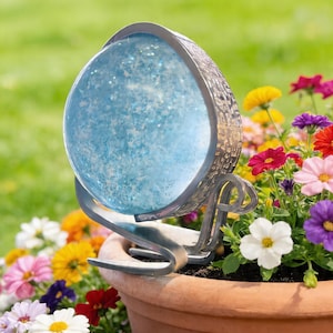 Puede incluir: Adorno decorativo de jardín al aire libre con un globo de cristal azul lleno de purpurina, sostenido en un marco de metal plateado. El adorno está colocado en una maceta de terracota rodeada de flores coloridas, con un fondo verde.