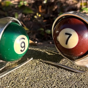 Puede incluir: Dos originales soportes hechos a mano para bolas de billar. Uno sostiene una bola verde con el número 9, el otro una bola marrón con el número 7. Cada bola está sujeta en un marco de metal plateado, que se asemeja a un caracol.