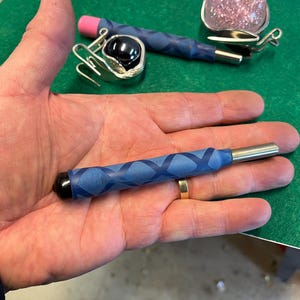 May include: A selection of jewellery tools, including a silver earring with a black stone, a silver earring with a pink stone, and a blue-taped tool with a black tip. The tools are displayed on a green surface.
