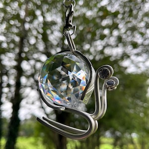 Puede incluir: Un adorno colgante con forma de caracol, fabricado con alambre de metal plateado y una esfera de cristal transparente facetado como concha. El caracol tiene dos antenas rematadas con pequeños brillantes transparentes y cuelga de una cadena.