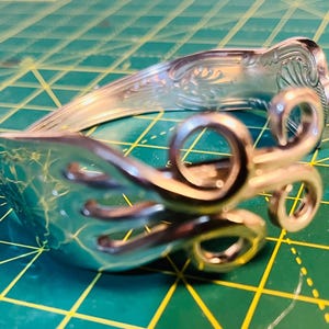 May include: A silver bracelet crafted from repurposed silverware. The bracelet features a fork head with tines and a decorative handle. The design includes scrollwork and a curved shape, showcasing a unique, handcrafted aesthetic.