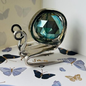 Puede incluir: Una figura decorativa de caracol plateado con una gran concha de cristal verde azulado facetada. El cuerpo del caracol es de metal curvado, y la base tiene la palabra "Say". La figura está colocada sobre una superficie decorada con ilustraciones de mariposas.