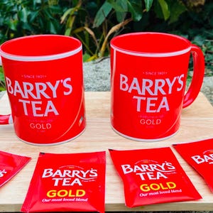Könnte beinhalten: Zwei rote Barry's Tea-Tassen und vier Teebeutel sind auf einer Holzoberfläche angeordnet. Die Tassen haben einen weißen Rand und das Barry's Tea-Logo. Die Teebeutel sind rot mit dem Barry's Tea Gold-Logo und Text.