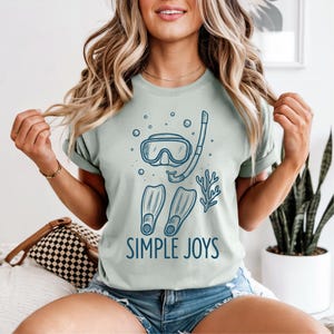 May include: A light green t-shirt featuring a blue illustration of snorkeling gear, including a mask, snorkel, and fins. The words "SIMPLE JOYS" are printed below the graphic in blue.