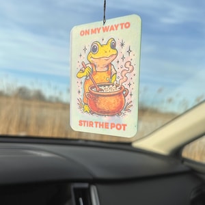 May include: Rectangular air freshener with a cartoon frog stirring a pot. The frog wears an orange apron and the text reads "ON MY WAY TO STIR THE POT". The background is a field and blue sky.