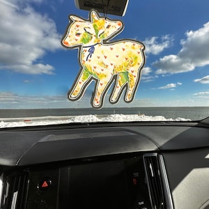 May include: A decorative car air freshener shaped like a two-headed deer. The deer is yellow with red heart accents and green details. It hangs from the rearview mirror against a blue sky with clouds.