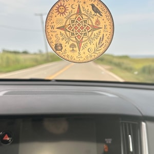 Puede incluir: Un ambientador de coche circular colgado del espejo retrovisor, con fondo beige y un diseño de brújula central. Muestra los puntos cardinales N, S, E, O, etiquetados como Aire, Tierra, Fuego y Agua, junto a ilustraciones de un sol, un cuervo, un caldero y un árbol.