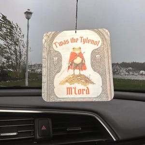 May include: Car air freshener with a whimsical design. The rectangular air freshener features a frog in a red cape holding a sword, with the text "T'was the Tylenol M'lord". The air freshener is hanging from a black string.