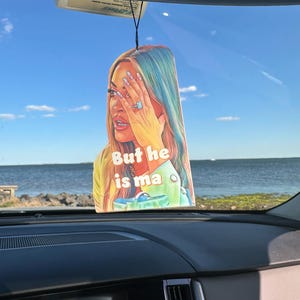 Può includere: Un deodorante per auto rettangolare con un'illustrazione colorata di una persona con capelli blu e verdi, che si copre il viso con la mano. Il testo "But he is ma" è visibile. Il deodorante è appeso a un filo nero.