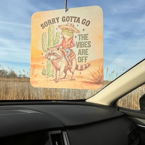 May include: Rectangular air freshener with a cartoon illustration of a frog in a cowboy hat riding a raccoon. The text reads "Sorry Gotta Go + The Vibes Are Off" with a desert landscape background.