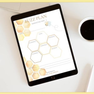 May include: A digital marketing plan displayed on a tablet. The "Buzz Plan" layout features honeycomb shapes for goal setting, audience analysis, and strategy. The tablet is on a white surface with a coffee cup and a pen.