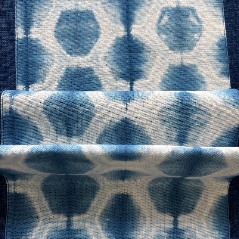 May include: Close-up of indigo blue and white fabric with a geometric pattern. The design features repeating hexagon shapes with a tie-dye effect. The fabric is folded, revealing layers of the patterned material.