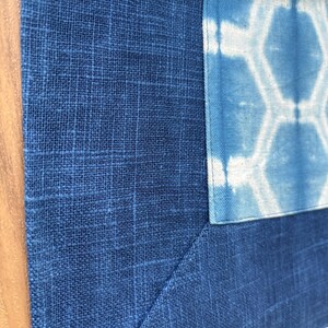 May include: Close-up of a textile piece featuring a patchwork design. The main fabric is a deep blue with a grid-like texture. A lighter blue section with a white geometric pattern is sewn into the corner, creating a visual contrast.