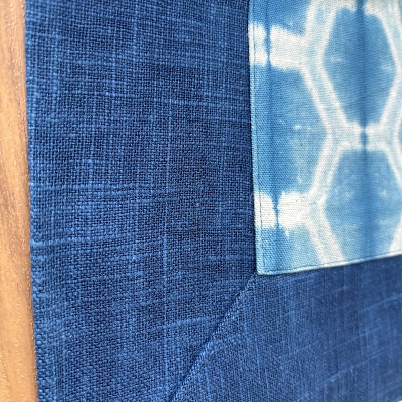 May include: Close-up of a textile piece featuring a patchwork design. The main fabric is a deep blue with a grid-like texture. A lighter blue section with a white geometric pattern is sewn into the corner, creating a visual contrast.