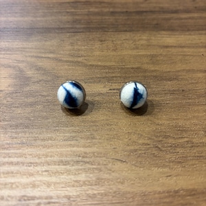 Può includere: Un paio di piccoli orecchini rotondi con un motivo tie-dye bianco e blu. Gli orecchini hanno una montatura in metallo argentato.