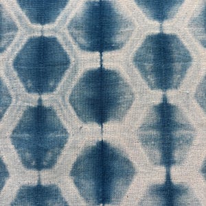 May include: Close-up of a textile with a repeating pattern of blue hexagonal shapes against a white background. The fabric appears to be hand-dyed, with variations in the blue color creating a textured effect. The design is geometric and symmetrical.