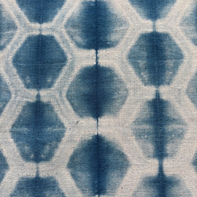May include: Close-up of a textile with a repeating pattern of blue hexagonal shapes against a white background. The fabric appears to be hand-dyed, with variations in the blue color creating a textured effect. The design is geometric and symmetrical.