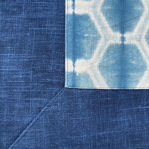 May include: Close-up of a section of blue fabric with a geometric pattern. The fabric has a textured appearance, with a darker blue section meeting a lighter blue section with a white geometric design. The edges are neatly sewn.