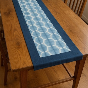 Może przedstawiać: Prostokątny bieżnik na stół z niebiesko-białym geometrycznym wzorem. Bieżnik jest obramowany jednolitym granatowym materiałem. Leży na drewnianym stole, widoczne są drewniane krzesła.