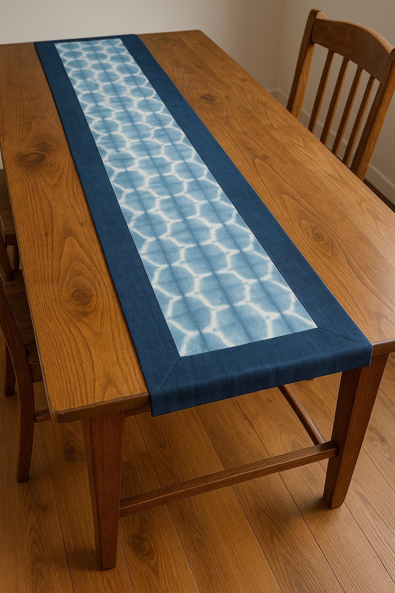 May include: A rectangular table runner with a blue and white geometric pattern. The runner is bordered with a solid navy blue fabric. The runner is placed on a wooden table, with wooden chairs visible.