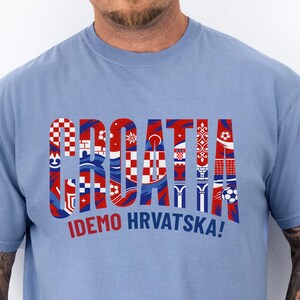 May include: A light blue t-shirt with the word "CROATIA" in a red, white, and blue design, with the text "IDEMO HRVATSKA!" below. The shirt is made of a soft material.
