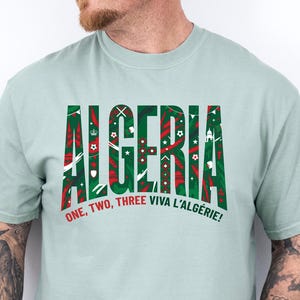 K&ouml;nnte beinhalten: Hellgr&uuml;nes T-Shirt mit dem Wort "ALGERIA" in Gr&uuml;n und Rot, mit dekorativen Mustern. Unter dem Wort steht der Satz "ONE, TWO, THREE VIVA L'ALG&Eacute;RIE!"