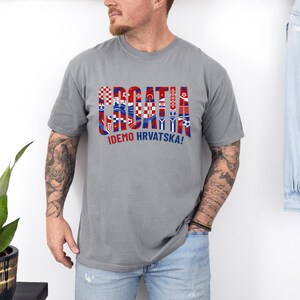May include: A gray t-shirt with the word "CROATIA" in a colorful, patterned design, with the text "IDEMO HRVATSKA!" below. The shirt is short-sleeved and appears to be made of a soft material.