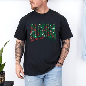 K&ouml;nnte beinhalten: Schwarzes T-Shirt mit dem Wort "ALASKA" in einem gr&uuml;nen und roten Muster. Darunter steht der Text "ONE, TWO, THREE, ALASKA". Das T-Shirt wird von einer Person getragen.