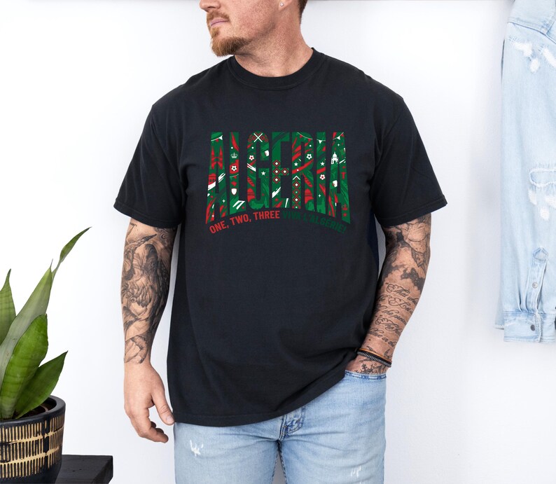 K&ouml;nnte beinhalten: Schwarzes T-Shirt mit dem Wort "ALASKA" in einem gr&uuml;nen und roten Muster. Darunter steht der Text "ONE, TWO, THREE, ALASKA". Das T-Shirt wird von einer Person getragen.
