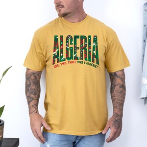 K&ouml;nnte beinhalten: Senfgelbes T-Shirt mit dem Wort "ALGERIA" in gro&szlig;en, gr&uuml;n und rot gemusterten Buchstaben. Darunter steht der Text "ONE, TWO, THREE VIVA L'ALG&Eacute;RIE!"
