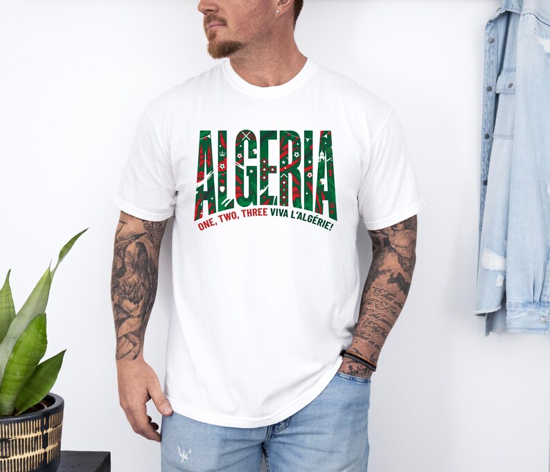 K&ouml;nnte beinhalten: Wei&szlig;es T-Shirt mit dem Wort "ALGERIA" in gr&uuml;nen und roten Blockbuchstaben. Das Shirt tr&auml;gt auch den Text "ONE, TWO, THREE VIVA L'ALG&Eacute;RIE!" in Rot. Das Shirt hat einen Rundhalsausschnitt.