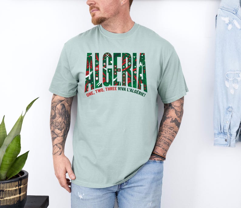 K&ouml;nnte beinhalten: Hellgr&uuml;nes T-Shirt mit dem Wort "ALGERIA" in Gr&uuml;n und Rot, mit dekorativen Mustern. Unter dem Wort steht der Satz "ONE, TWO, THREE VIVA L'ALG&Eacute;RIE!"