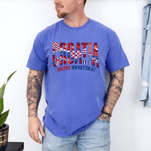 May include: A blue t-shirt with the word "CROATIA" in a multi-colored design, incorporating Croatian flag elements. Below the word is the phrase "IDEMO HRVATSKA!" in white. The shirt is worn by a person.