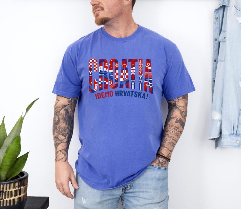 May include: A blue t-shirt with the word "CROATIA" in a multi-colored design, incorporating Croatian flag elements. Below the word is the phrase "IDEMO HRVATSKA!" in white. The shirt is worn by a person.