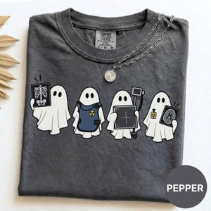 May include: A charcoal gray t-shirt featuring a graphic of four white ghosts. The first ghost holds an X-ray of a ribcage, the second wears a blue radiation vest, the third holds a medical imaging device, and the fourth holds a brain scan.