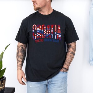 May include: Black t-shirt with a Croatian flag design. The word "CROATIA" is displayed in a stylized font, incorporating red, white, and blue elements. Below the word is the phrase "IDEMO HRVATSKA!"