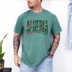 K&ouml;nnte beinhalten: Ein gr&uuml;nes T-Shirt mit dem Wort "ALGERIA" in einer dekorativen Schriftart, gef&uuml;llt mit roten, wei&szlig;en und gr&uuml;nen Mustern. Darunter steht der Satz "ONE, TWO, THREE VIVA L'ALGERIE!" in Rot und Wei&szlig;. Das T-Shirt wird von einer Person getragen.